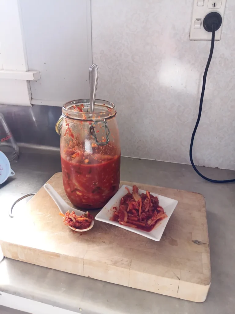 Easy kimchi recipe: a half-gallon jar of homemade kimchi on a chopping board, with a spoonful and a small plate of the finished kimchi beside it