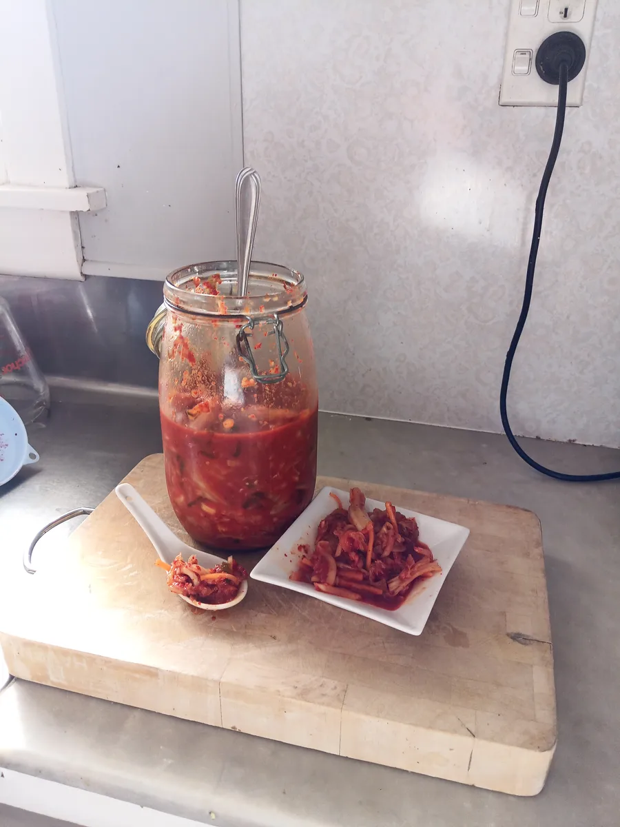 Easy kimchi recipe: a half-gallon jar of homemade kimchi on a chopping board, with a spoonful and a small plate of the finished kimchi beside it