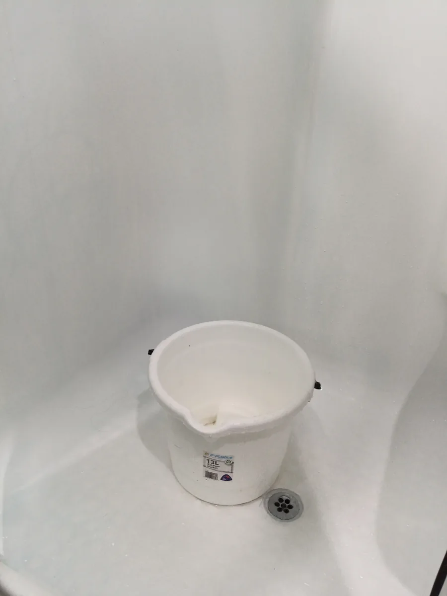A white plastic bucket on a shower floor next to the drain.