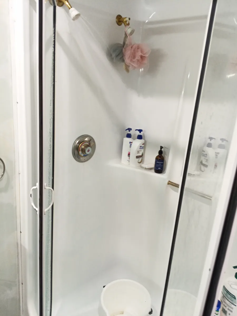 A white bucket on the floor of a shower stall under the running showerhead.